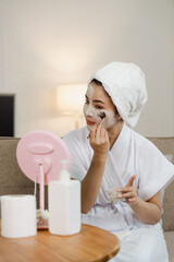 Young pretty asian woman with facial mask sitting on sofa puts nourishing mask on skin for...