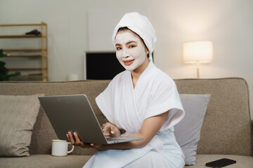 Young pretty asian woman with facial mask sitting on sofa puts nourishing mask on skin for rejuvenating and eliminating defects. Acne. Smile.