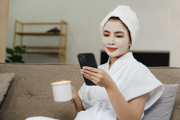 Young pretty asian woman with facial mask sitting on sofa puts nourishing mask on skin for rejuvenating and eliminating defects. Acne. Smile.