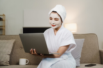 Young pretty asian woman with facial mask sitting on sofa puts nourishing mask on skin for...