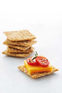 Cracker topped with Cheddar Cheese, Tomato Slices and Herbs with Stack of Crackers in Background on White Background, Studio Shot