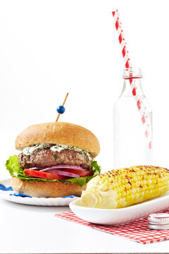 Greek style burger with spiced corn on the cob and bottle of soda with striped straws, studio shot on white background