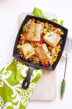 Mahi-Mahi on couscous in a cast-iron skillet with a green patterned napkin, studio shot on white background