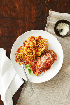 Plate of chicken parmesan with spaghetti and tomato sauce and a side dish of cheese with a fork and napkin on a wooden background