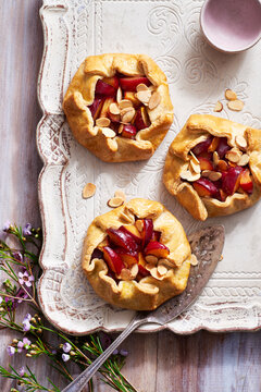 Three plum almond galettes on a decorative, rustic serving tray with pie server and purple flowers