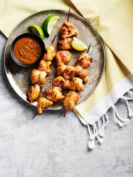 Chicken satay skewers with dipping sauce and lime wedges on a metal tray on striped fabric