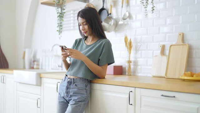 Happy young asian woman relaxing at home. Asia female standing at counter kitchen and using mobile smartphone