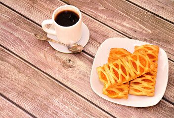 A cup of hot coffee on a saucer with a spoon and a plate with three puff buns with fruit jam on a wooden table.