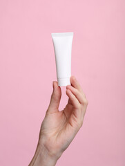 Woman's hand holds white tube of cream on pink background