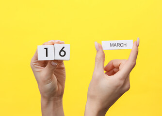 Woman's hands hold wooden block calendar with the date march 16 on yellow background
