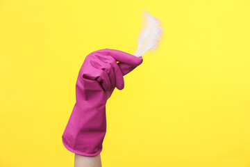 Hand in purple rubber cleaning glove holding feather on a yellow background. House cleaning and housekeeping concept