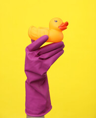Hand in purple rubber cleaning glove holding rubber duck on a yellow background. House cleaning and housekeeping concept