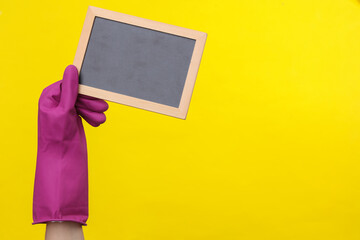 Hand in purple rubber cleaning glove holding empty chalk board on a yellow background. House cleaning and housekeeping concept