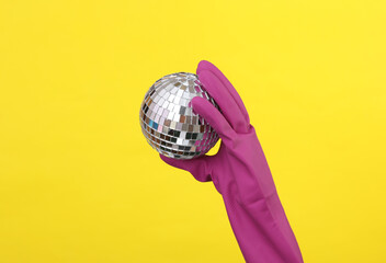 Hand in purple rubber cleaning glove holding disco ball on yellow background. House cleaning and housekeeping concept. Party concept