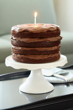 Layered Chocolate Birthday Cake with Lit Candle on Cake Stand on Coffee Table