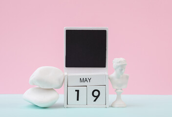 Block wooden calendar with the date may 19 and white pebbles stack with Venus bust on pastel background.