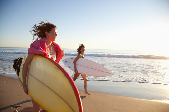Young Women Running on Beach Holding Surf Boards, Zuma Beach, California, USA