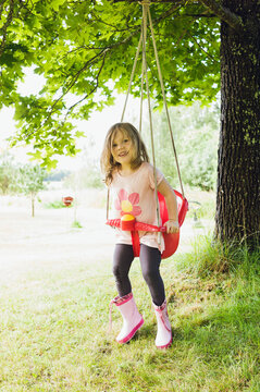 3 year old girl in rubber boots sitting in red swing in back yard, looking at camera and smiling, Sweden