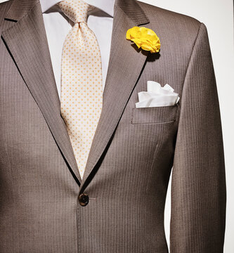 Detail of a light brown suit jacket with cream colored shirt and white handkerchief, yellow patterned necktie and yellow flower