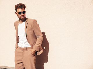Handsome confident stylish hipster lambersexual model.Sexy modern man dressed in elegant beige suit. Fashion male posing in the street in city at sunset. In sunglasses. Near wall - obrazy, fototapety, plakaty
