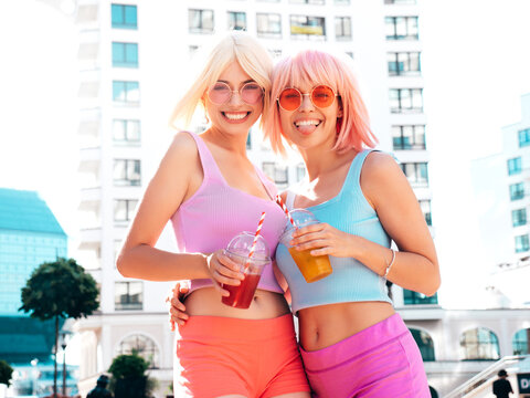 Two young beautiful smiling hipster female in trendy summer clothes. Sexy carefree women posing in the street in wigs. Positive funny models drinking fresh cocktail smoothie drink in plastic cup
