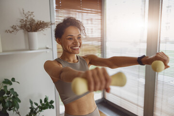 Fototapeta na wymiar Smiling woman with dumbbells doing exercising at home and working on arms strength