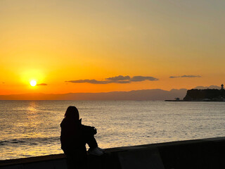 夕焼けの海を膝を抱えて眺める女性のシルエットと江の島