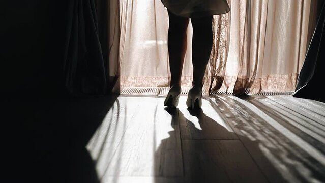 The silhouette of the legs of a girl standing in front of a bright window leading to the balcony. On the girl's feet are shoes