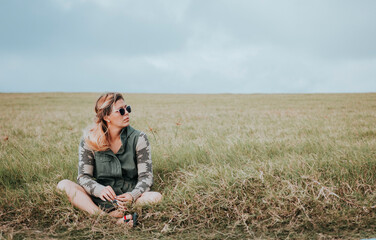 Naklejka na ściany i meble Portrait of girl in glasses sitting cross legged in the field. Relaxed teen girl sitting in the field looking to the side. Concept of young woman sitting and relaxing in the field