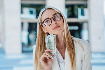 Pensive blonde caucasian woman in glasses, white suite holds US dollar banknotes looks aside, upset by profit, stands against blurry buildings. Entrepreneur unhappy loosed money, small salary.
