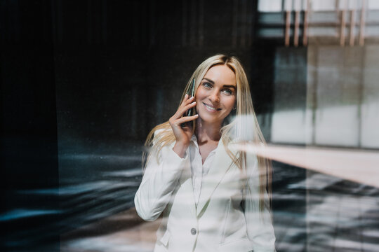 Beautiful blonde businesswoman in white suite talking by phone, broad smiling, looking up through the window on sunny day, excited by good news. Happy Scandinavian woman at work, loving her job.