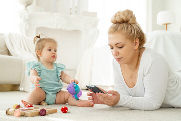 Obraz na płótnie Canvas Young mom using smartphone while playing with baby. Lack of live communication, Addiction on gadgets. Social media, watch video, Woman search for products for children in Internet. 