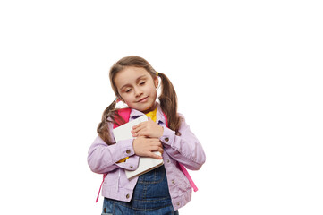 Caucasian smart little girl with ponytails, wearing purple jacket and denim overalls, gently hugging her textbook, posing with her eyes closed over white background. Schoolgirl. Pupil. Smart student