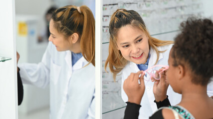 Fototapeta premium Smiling female optician helping child girl choosing the most appropriate eyeglasses. Eyesight and vision concept