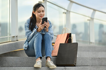Pleasant asian woman using mobile phone while sitting on rooftop stair with shopping bags