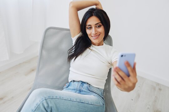 A woman with a phone sits on a chair in jeans and a T-shirt and takes pictures of herself on her phone camera, a blogger poses and takes selfies