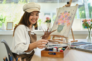 Pleasant female artist in apron with palette and brush painting on canvas at art studio. Education,...