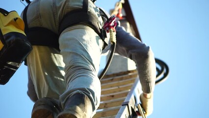 Skilled electrician in helmet fixes wires standing on ladder near high pole against blue sky on summer day backside view. Electrical service and mounting on the pole. Slow motion