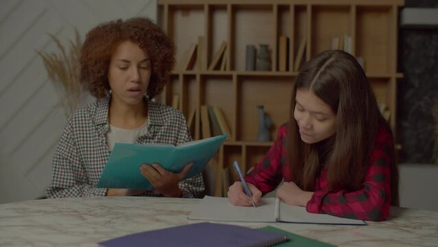 Caring charming African American mother with tutorial reading school assignment, helping smart multicultural teenager daughter to do homework while family studying together in domestic room.