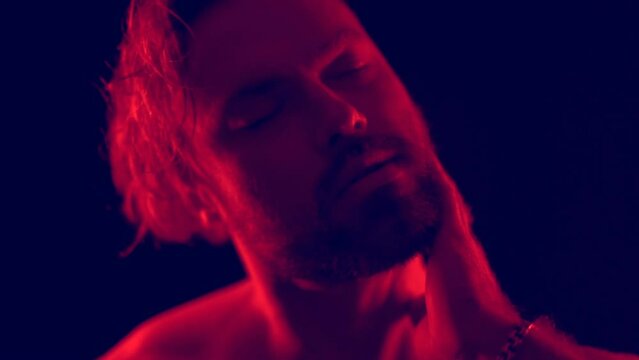 Dark video closeup portrait of a young blonde bearded caucasian man with red light in slow motion. Fashion trendy studio videoclip gogo dancer isolated with black background