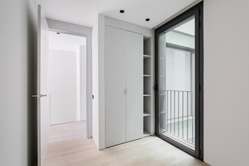 Modern style entrance hall interior, neutral white color corridor with wooden light parquet floor, sliding wardrobe and glass door in black aluminum frame with access to the street.
