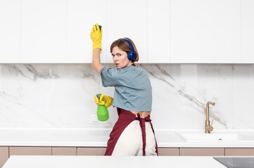 Young attractive housewife in headphones and apron listening to music and dancing during household...