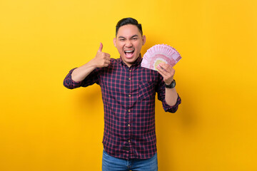 Fototapeta na wymiar Excited young Asian man holding money banknotes and gesturing thumbs up, celebrating financial success isolated over yellow background. Profit and wealth concept