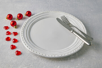An empty white ceramic plate with cutlery surrounded by red christmas balls and hearts on a light concrete table