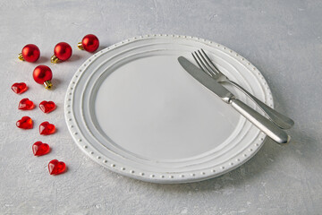 An empty white ceramic plate with cutlery surrounded by red christmas balls and hearts on a light...