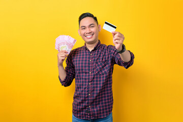 Happy young Asian man holding money banknotes and credit card isolated over yellow background. Easy...