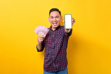 Excited young Asian man holding mobile phone with blank screen and money banknotes isolated over yellow background. Easy online money transfer concept