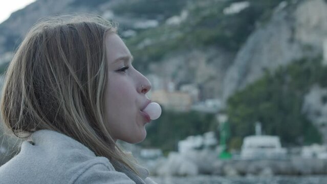 Side Portrait Of Beautiful Young Woman Blowing Bubble Gum With Amalfi Coast In Backdrop. closeup