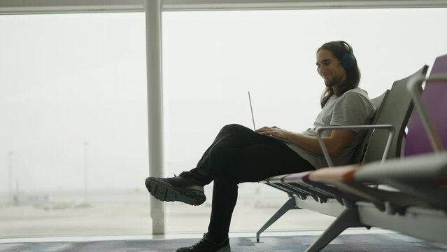 Adult digital nomad man looking at the camera and smiling while using his laptop sit on an airport
