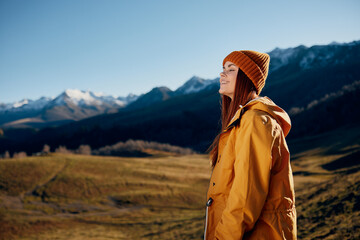 Woman standing beautifully on a hill smile with teeth in the mountains in the autumn in a yellow raincoat and jeans happy sunset trip to hike the mountains in the snow, freedom lifestyle 